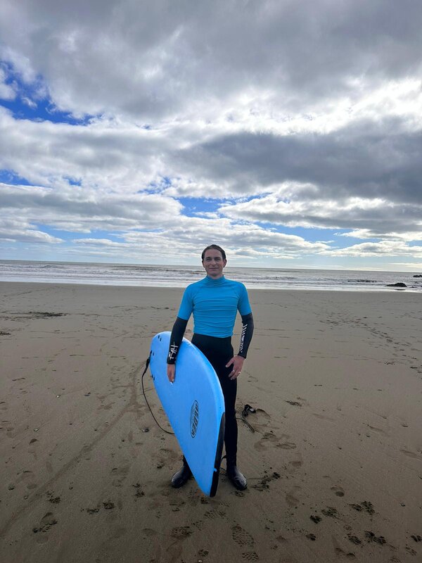 Ein junger Mann in einem Neoprenanzug und blauem Shirt mit einem Surfboard am Strand.
