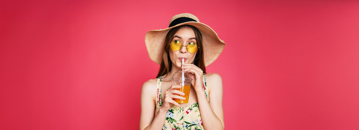 Junge Frau in floralem Sommerkleid und breitem Strohhut, die einen kalten Eistee im Plastikbecher mit Trinkhalm hält, vor kräftig pinkem Studiohintergrund, sommerlich-leichtes Stimmungsmotiv