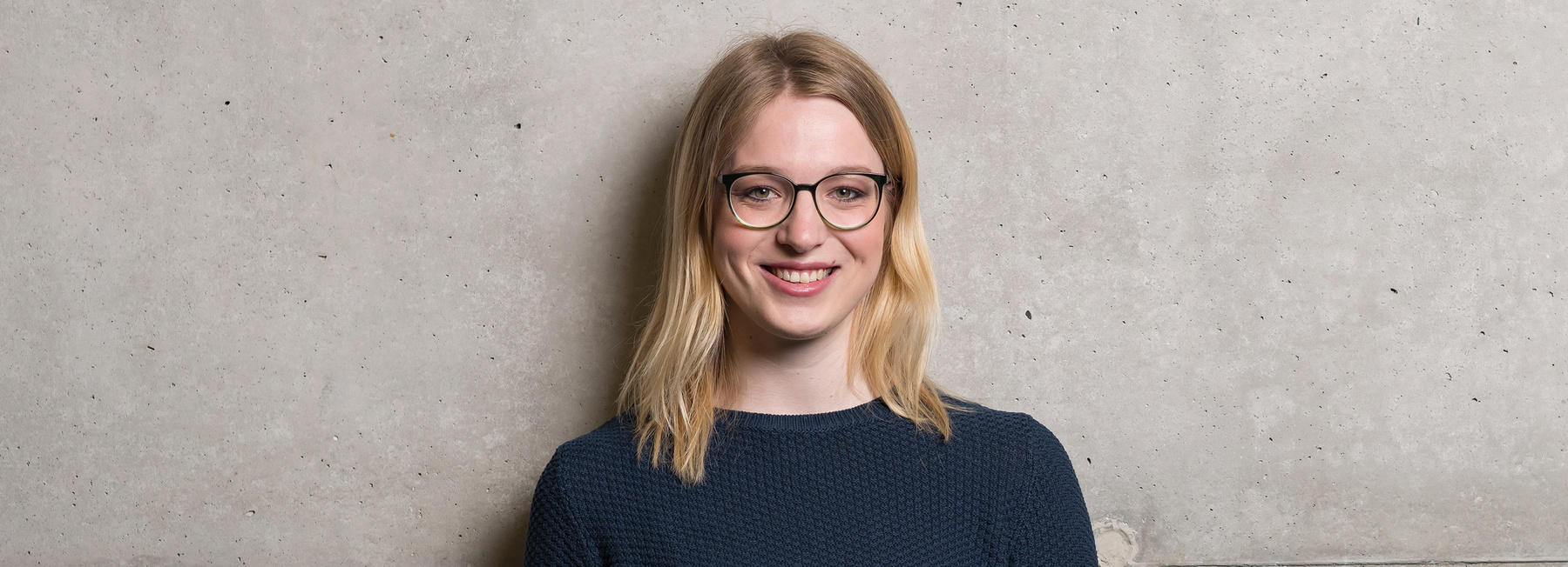 BA_ENM_VZ | © FH Kufstein Tirol Eine Frau steht mit dem Rücken zu einer grauen Wand und blickt lächelnd in die Kamera. | © FH Kufstein Tirol
