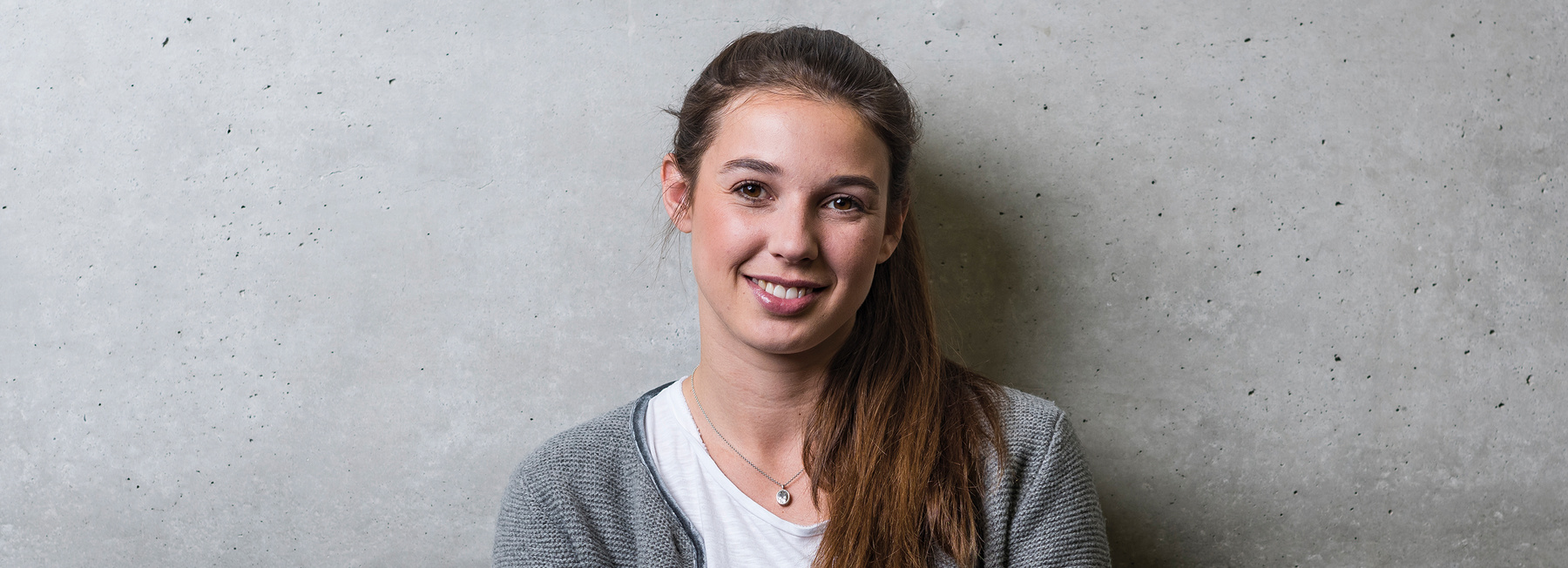 MA_ENM_BB | © FH Kufstein Tirol Eine Frau steht mit dem Rücken zu einer grauen Wand und blickt lächelnd in die Kamera. | © FH Kufstein Tirol