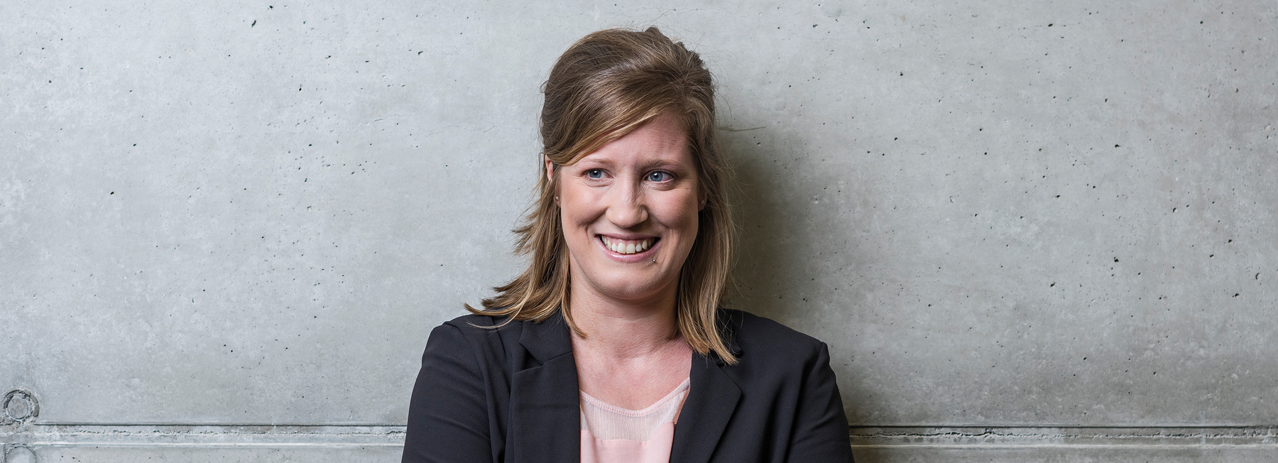 MA_DIM_BB | © FH Kufstein Tirol Eine Frau steht mit dem Rücken zu einer grauen Wand und blickt lächelnd in die Kamera. | © FH Kufstein Tirol