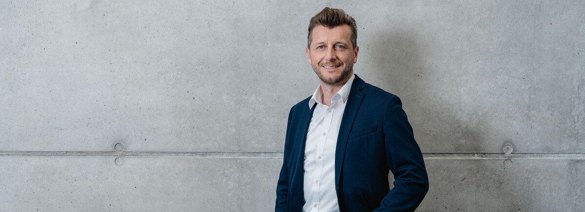 Über_die_International_Business_School_Sektion_3 | © FH Kufstein Tirol Ein Mann steht vor einer Betonwand und blickt lächelnd in die Kamera. | © FH Kufstein Tirol